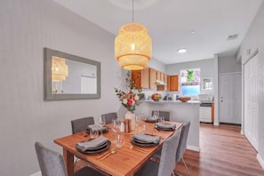 A dining area with a wooden table set for four, a hanging rattan light fixture, and a kitchen in the background.