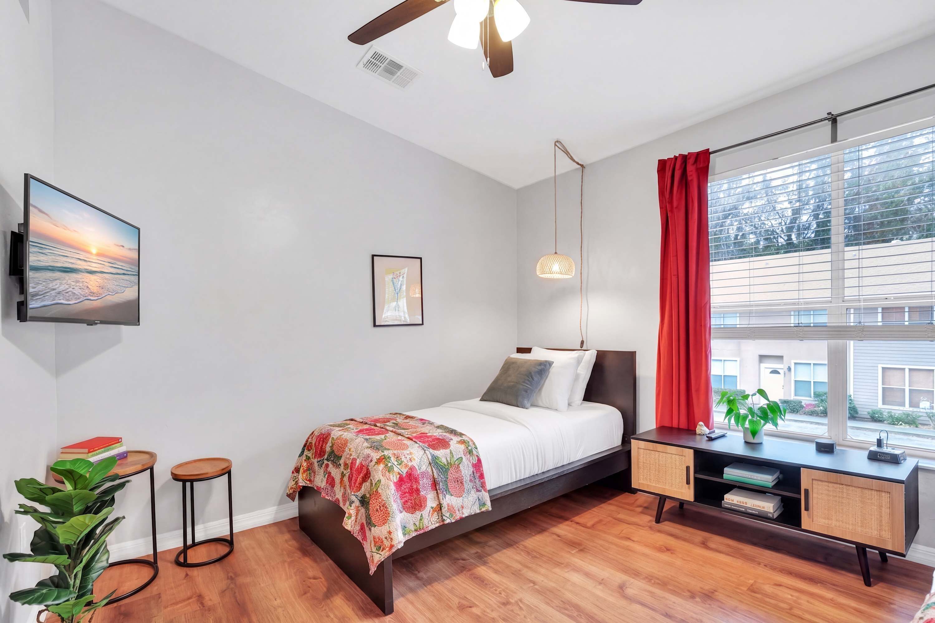 A bedroom with a bed, a desk, a TV, a window with curtains, and a plant.