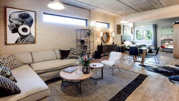 Una sala moderna con sofá, mesa de centro y sillas. Hay una zona de comedor con mesa y sillas. La habitación tiene un ventanal grande y un techo con vigas a la vista.