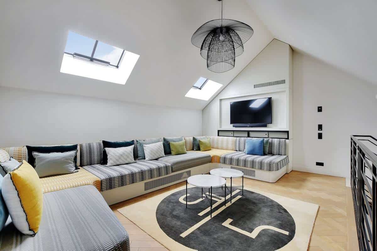 A modern loft-style living room with a large sectional sofa, a flat-screen TV mounted on the wall, skylights, and a unique black and white patterned rug.