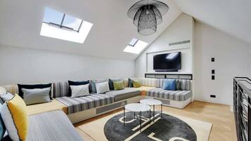 A modern loft-style living room with a large sectional sofa, a flat-screen TV mounted on the wall, skylights, and a unique black and white patterned rug.