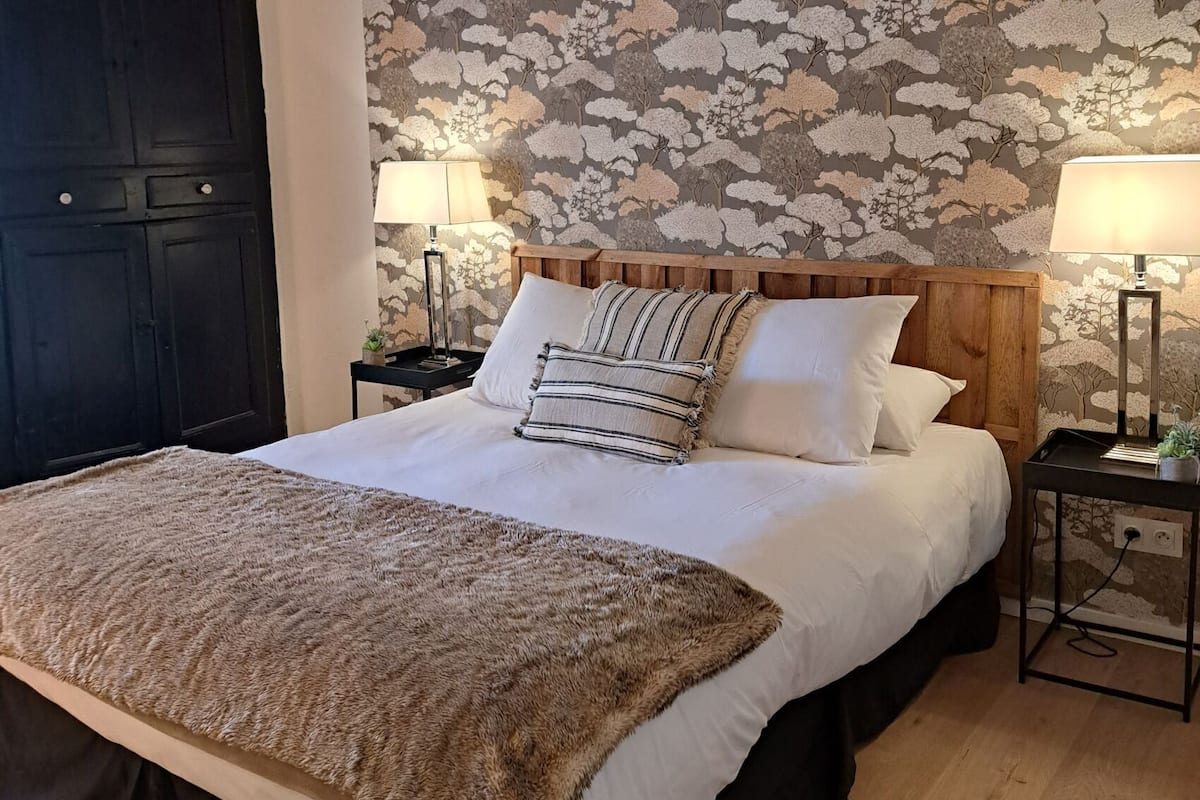A bedroom with a large bed, wooden headboard, floral wallpaper, two bedside tables with lamps, and a dark door.