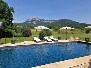 Zona de piscina con sillones de descanso y sombrillas, rodeada de vegetación y vista a la montaña.