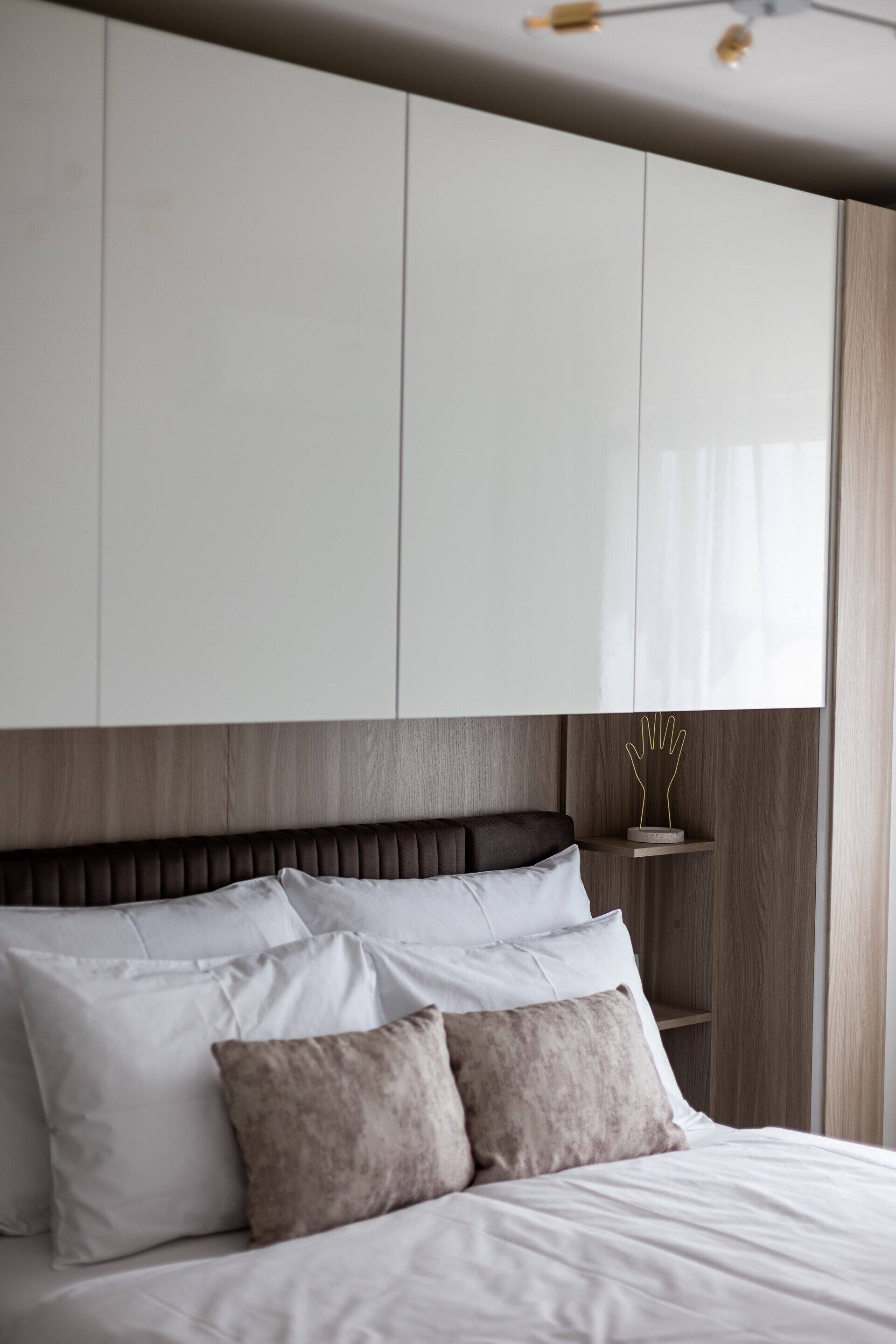 Une chambre à coucher avec un lit, des oreillers blancs et bruns, une tête de lit en bois, une armoire blanche et une petite étagère sur laquelle est posé un objet décoratif.