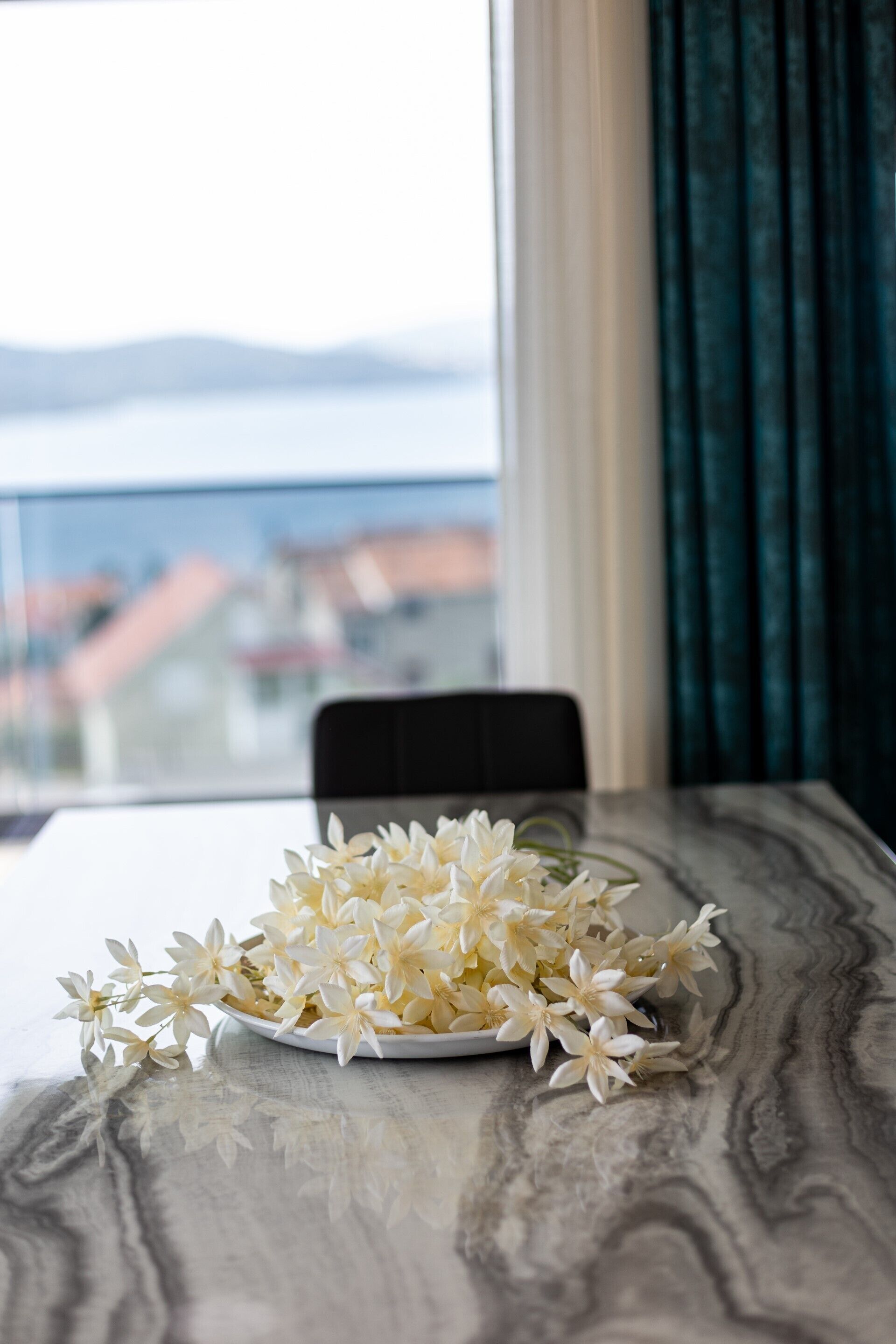 Ein Tisch mit einer weißen Teller mit weißen Blüten, ein Stuhl und Blick auf Gebäude durch ein Fenster.