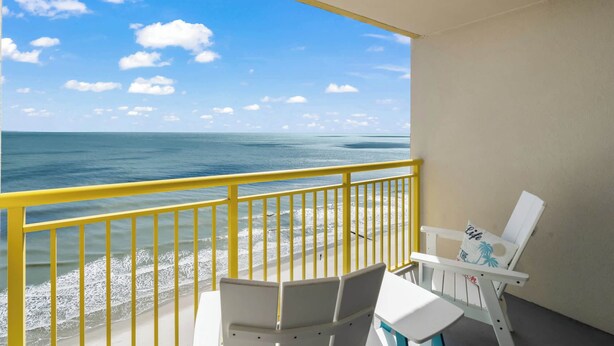 Een balkon met een gele balustrade, witte stoelen en een tafel, met uitzicht op een strand met helderblauw water en een zandstrand onder een lichtbewolkte lucht.