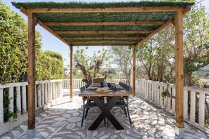 Una pérgola de madera al aire libre con una mesa lista para comer, rodeada de baldosas de piedra y barandas blancas.