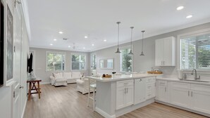 A modern kitchen with white cabinets, a central island, pendant lights, and a dining area with a white sofa and chairs. - Isla Guava: Boutique Resort, FREE Boat Dockage Sept-Oct Only! (Islamorada)
