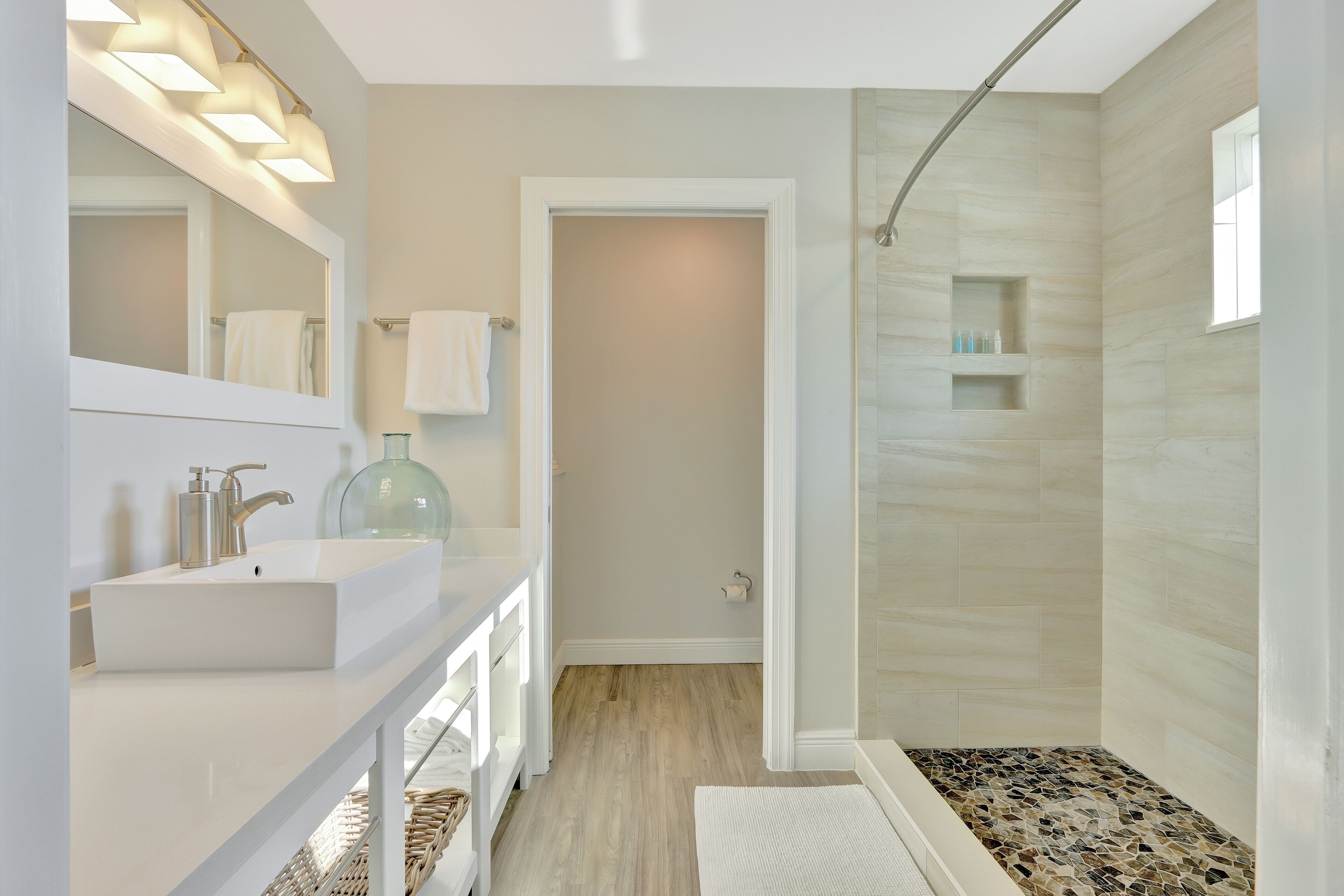 A modern bathroom with a white vanity, a large mirror, a glass jar, and a shower with a patterned floor.