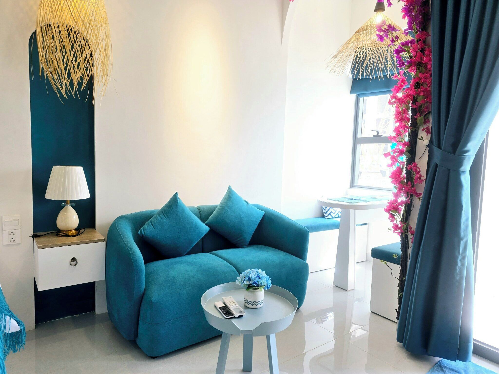 Un salon moderne avec un canapé de couleur turquoise, une petite table blanche ornée d’un vase et de télécommandes, une lampe posée sur une table d’appoint en bois, et une fenêtre avec des rideaux.