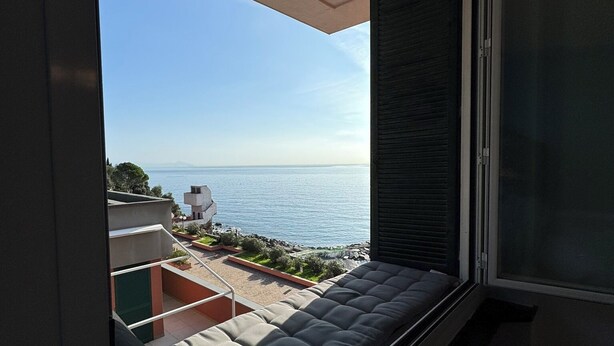 A balcony with a cushioned seating area, a view of the sea, and a building with a modern design. - La Soluzione Perfetta (Arenzano)