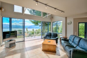 A spacious living room with a large TV, leather sofas, a wooden coffee table, and a balcony with a scenic mountain view. - Tawhiri - stunning views in paradise (Wānaka)