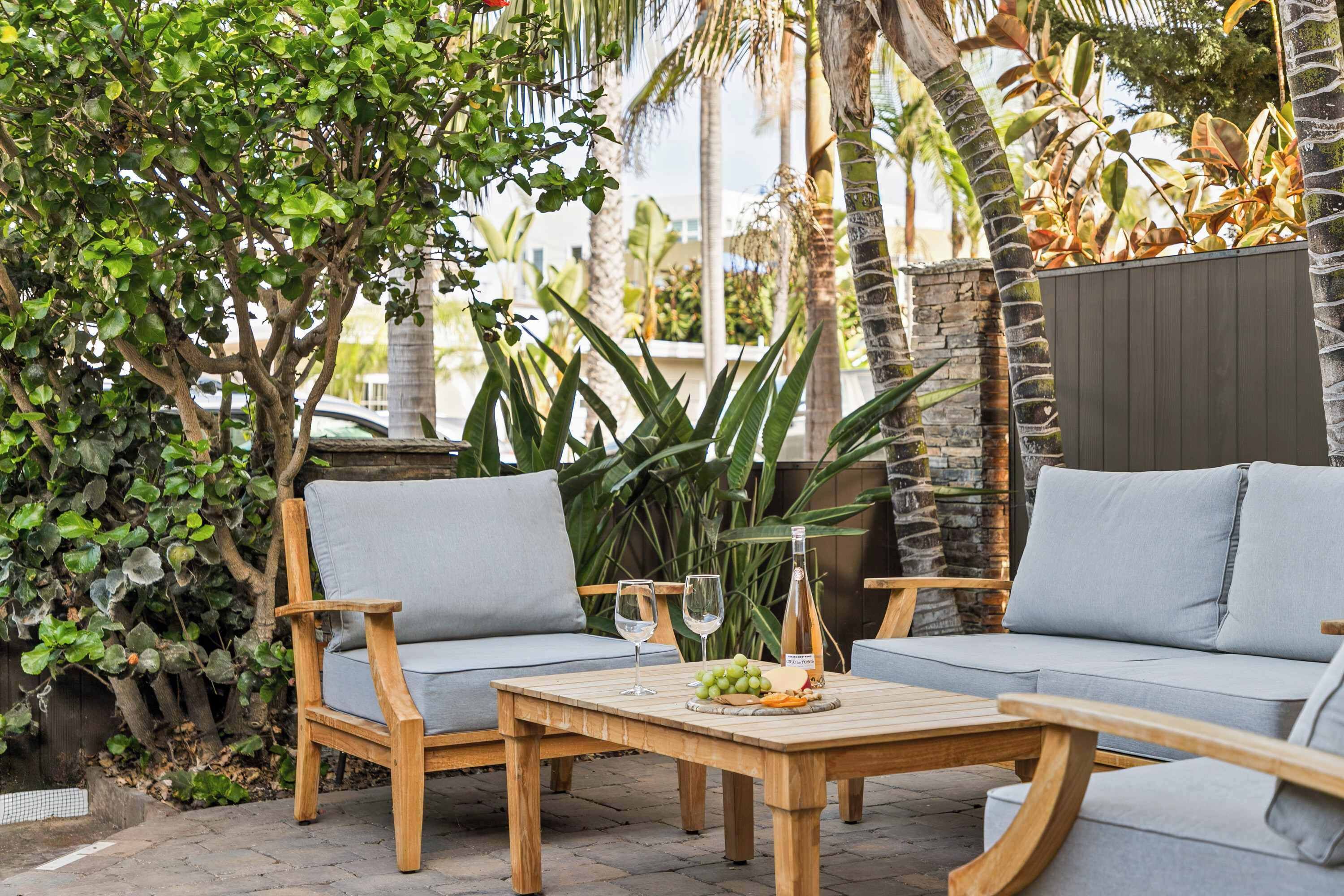 An outdoor patio with wooden furniture, a table with a bottle and grapes, and lush greenery.