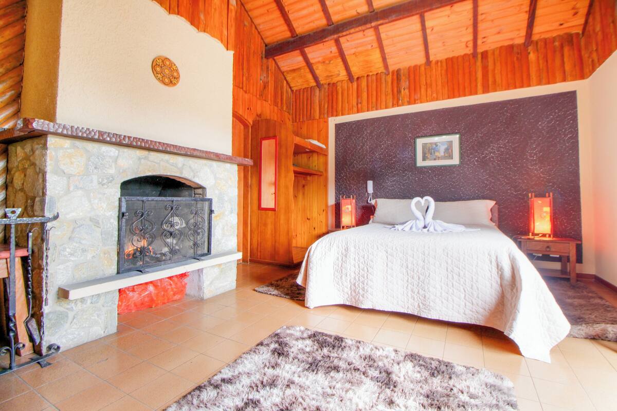 Quarto com lareira de pedra, cama com colcha branca e teto de madeira.