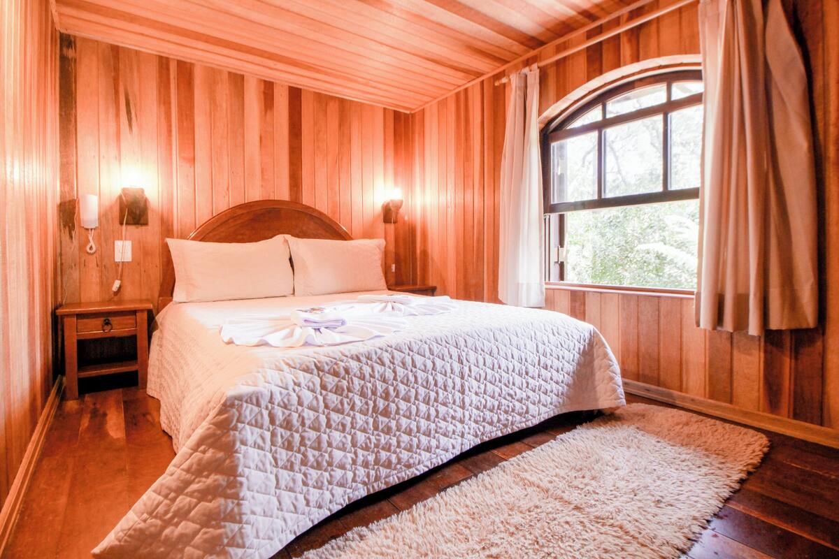 Quarto com painéis de madeira, cama, mesas de cabeceira e janela com cortinas.