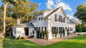 A two-story house with a covered porch, a stone wall, and a well-maintained lawn. - Stylish Cape Escape on Bank St RPAM070 (Harwich)