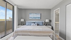 A bedroom with a large bed, two bedside tables, a bench, a sliding glass door to a balcony, and a painting of the sea on the wall. - ~ Oceanfront Oasis ~  Private Condo ~ 3 Bedroom (Myrtle Beach)