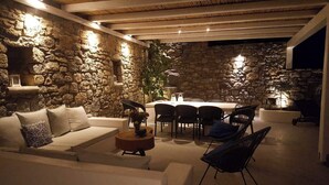 Un patio exterior con paredes de piedra, un sofá blanco, una mesa de comedor con sillas y una pequeña mesa de madera con un jarrón.