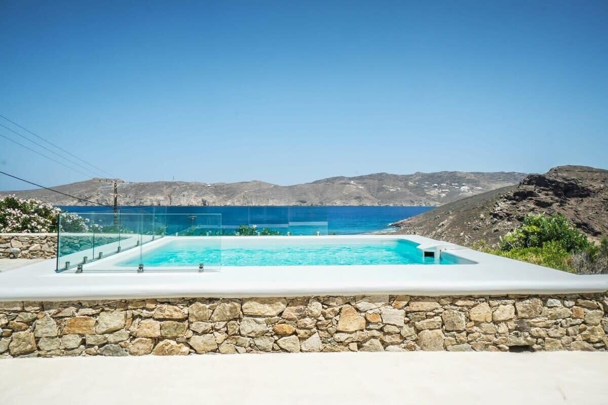 Ein rechteckiger Swimmingpool mit klarem, blauem Wasser, umgeben von einer Steinmauer, vor einer Kulisse aus felsigen Hügeln und einem wolkenlosen Himmel.