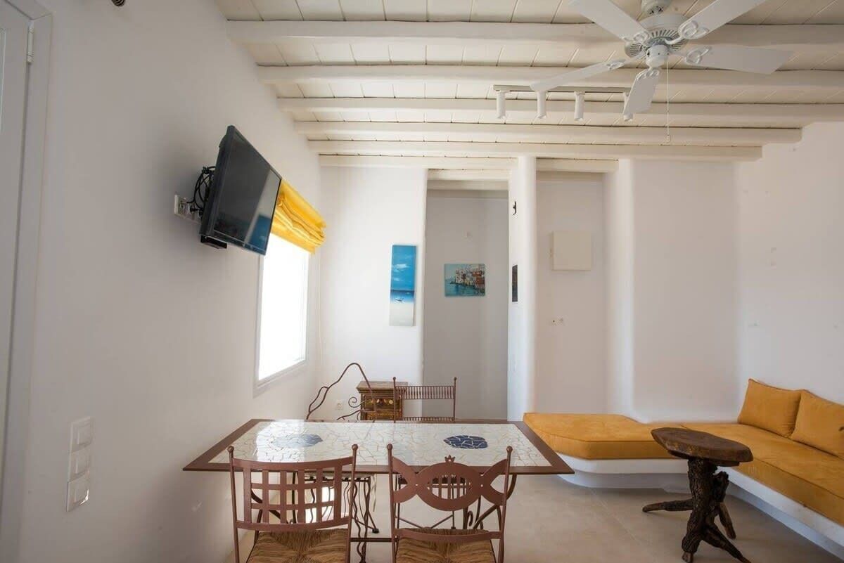 A dining area with a table and chairs, a sofa, a TV mounted on the wall, and a ceiling fan.