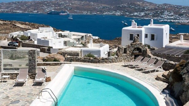 A pool area with white buildings, a clear blue sky, and a view of the sea and distant mountains.