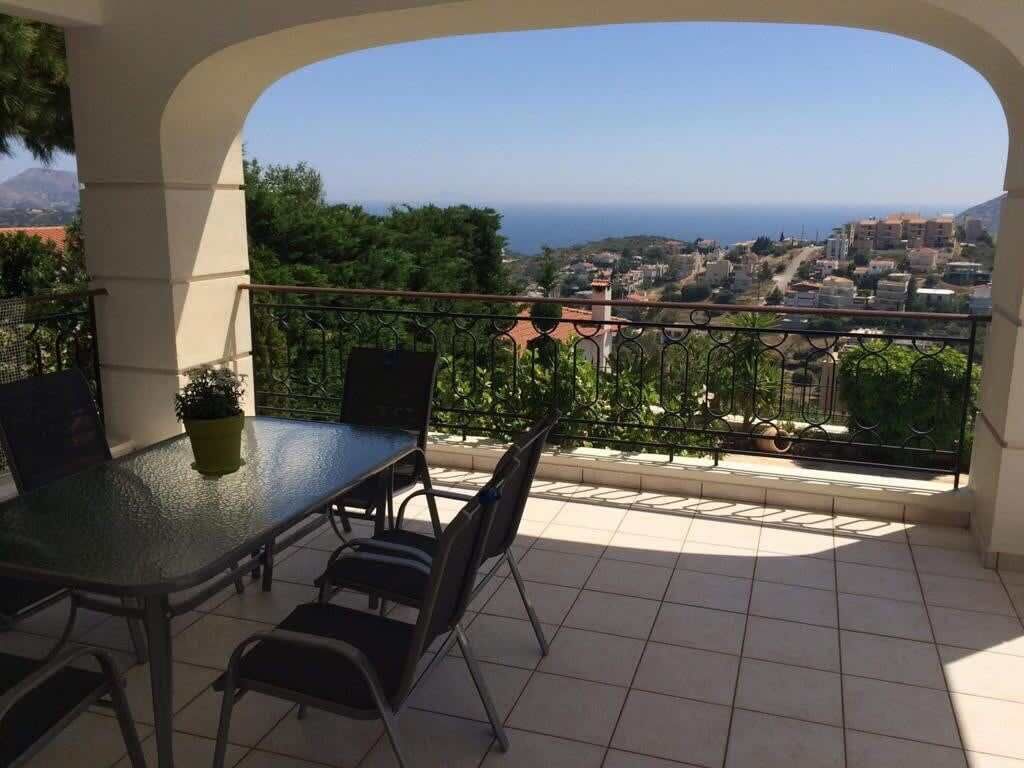 A balcony with a glass table and chairs, offering a view of a coastal town and the sea.