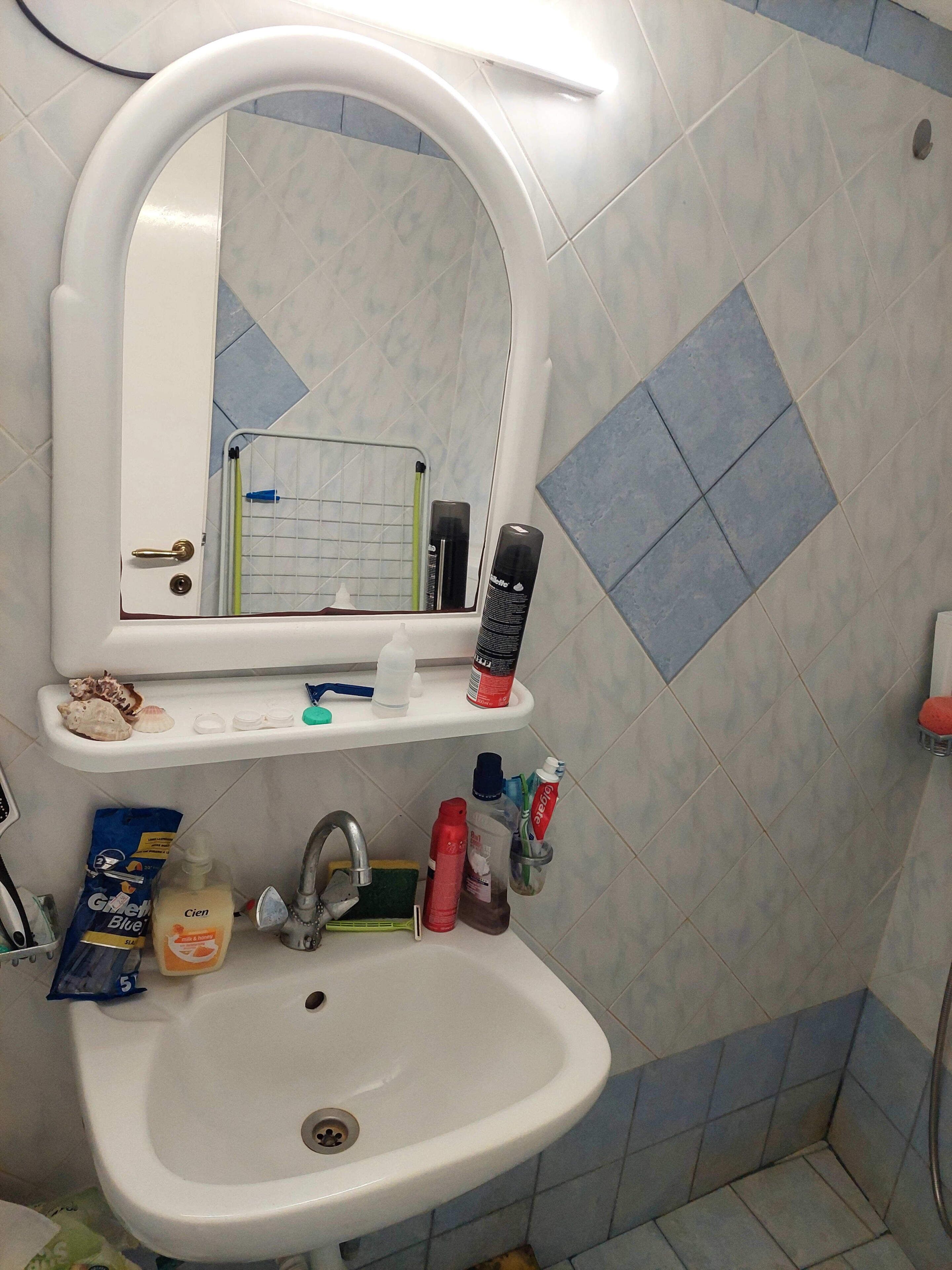 Um banheiro com uma pia branca, um espelho acima e uma prateleira com artigos de higiene pessoal.