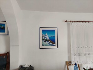A room with a framed picture of a coastal building, a wooden chair, a small table, and a curtain.