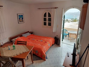 A bedroom with a bed, a table with chairs, a TV, and a view of the sea through an open door.