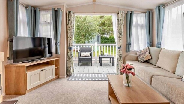A cozy living room with a TV on a wooden stand, a sofa, a coffee table, and a view of a deck and greenery outside.