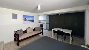 Un salon moderne avec un grand lit, un canapé et un coin repas comprenant une table et des chaises. Au fond, on aperçoit une cuisine avec des meubles sombres et un réfrigérateur.