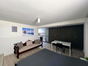 Ein modernes Wohnzimmer mit einem großen Bett, einer Couch und einem Essbereich mit Tisch und Stühlen. Im Hintergrund ist eine Küche mit dunklen Schränken und einem Kühlschrank zu sehen.