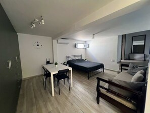 A compact room with a bed, a sofa, a dining table with chairs, and an air conditioning unit. - Charming Getaway in the Heart of Emporio Santorini (Santorini)