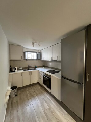 Eine kleine Küche mit weißen Schränken, einem Edelstahlkühlschrank, einem eingebauten Backofen und einem Fenster über der Spüle.