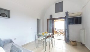 A modern living room with a dining area, a grey sofa, a TV mounted on the wall, and a large window. - Modern Naya Escape in Santorini with Captivating Sea Views (Santorini)