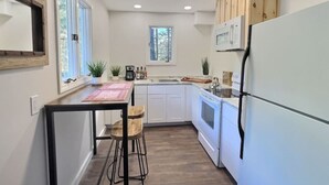 A compact kitchen with white cabinets, a white refrigerator, a microwave, and a stove. There is a window with vertical blinds, a small sink, and a table with two stools. - BearHaus1- Central Ac, Firepit, pool, gym (Killington)