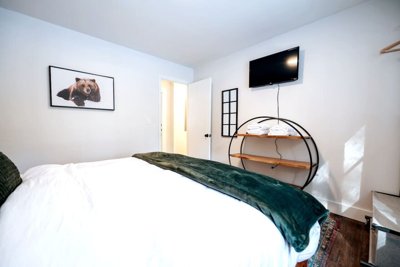 Une chambre à coucher avec un lit, un téléviseur fixé au mur, un tableau encadré accroché au mur et une étagère avec des serviettes.