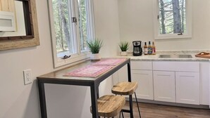 A kitchen with white cabinets, a wooden table with two stools, a window with a view of trees, and a coffee maker on the counter. - BearHaus1- Central Ac, Firepit, pool, gym (Killington)