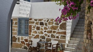A stone-walled outdoor seating area with a table and chairs, a window with blue shutters, and a staircase leading up. - Relaxing Patmos Studio by the Aegean No 3 (Patmos)