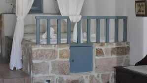 A room with a stone wall bed, a blue headboard, a white canopy, a small blue cabinet, and a white table with chairs. - Relaxing Patmos Studio by the Aegean No 3 (Patmos)