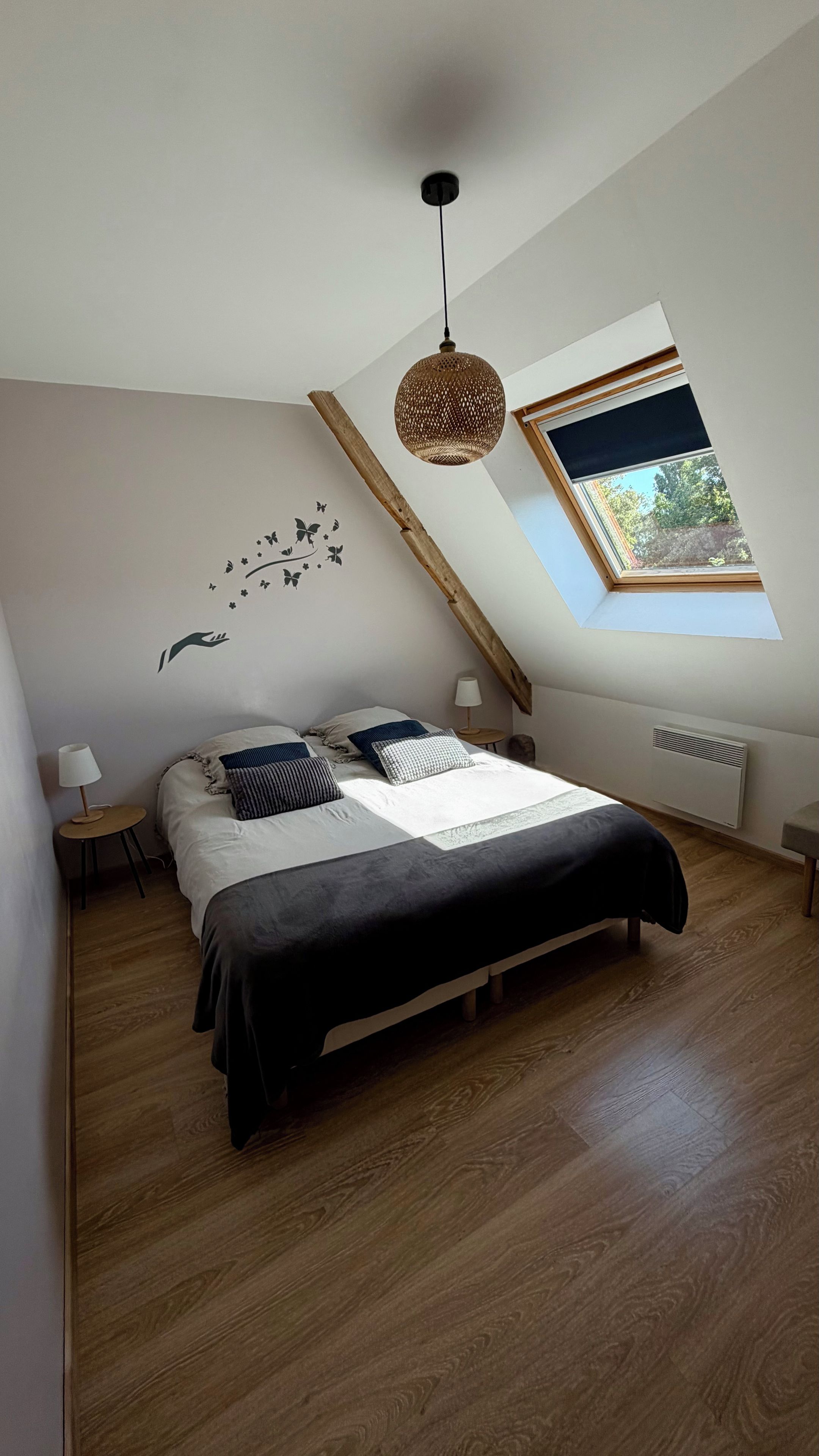 Een slaapkamer met een schuine plafond, een bed met veel kussens, een wandlamp, een hanglamp en een raam met uitzicht op groen.