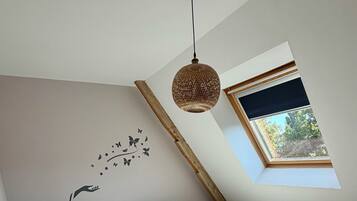 Een slaapkamer met een schuine plafond, een bed met veel kussens, een wandlamp, een hanglamp en een raam met uitzicht op groen.