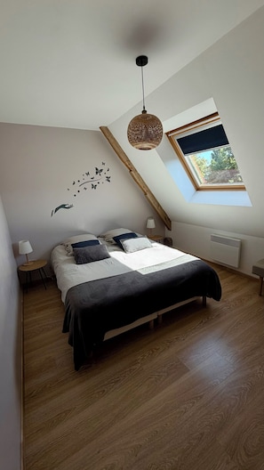 Une chambre avec un plafond en pente, un lit avec de nombreux oreillers, une lampe fixée au mur, un luminaire suspendu et une fenêtre donnant sur un paysage verdoyant.