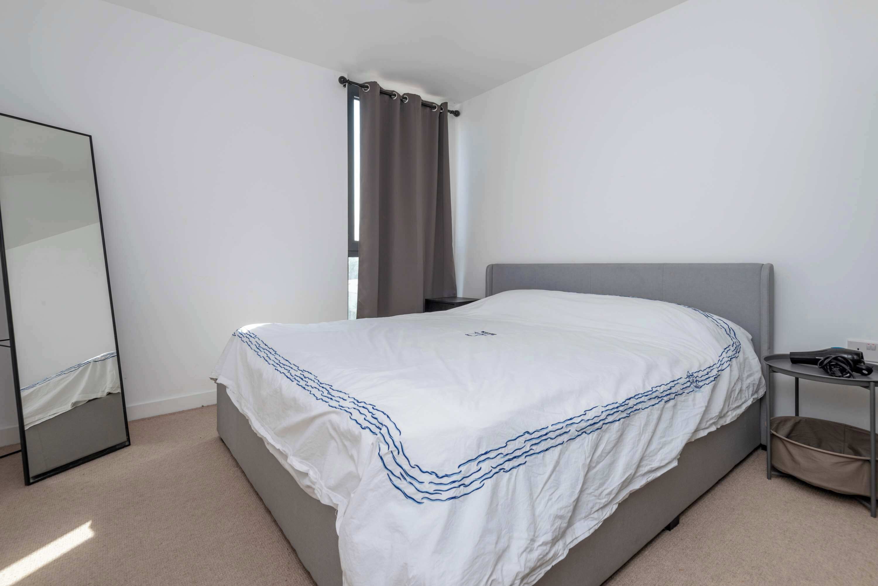 A bedroom with a bed, a bedside table, a mirror, and a window with curtains.