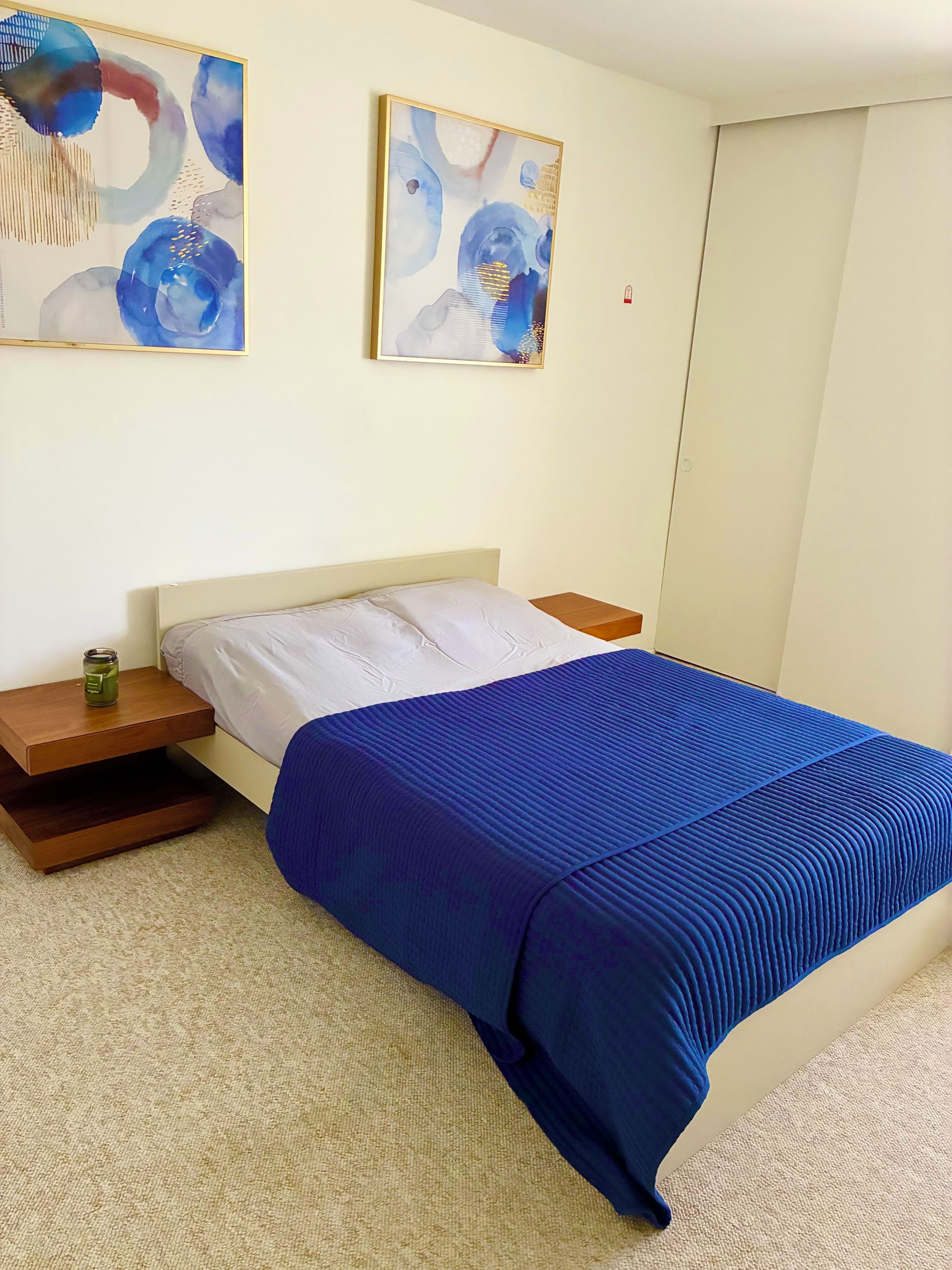 Een slaapkamer met een bed, twee nachtkastjes, een houten bijzettafeltje en twee abstracte schilderijen aan de muur.