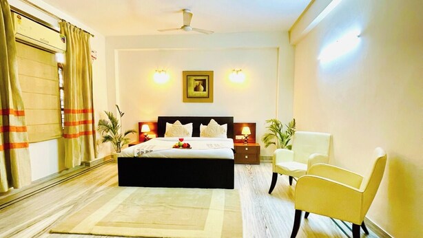 A hotel room with a bed, a ceiling fan, a nightstand, a chair, and a large window with curtains. - BedChambers Serviced Apartments -Artemis (Gurugram)