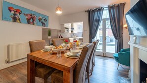 Un coin repas avec une table en bois dressée pour quatre personnes, un grand tableau accroché au mur et une vue sur l’extérieur par une porte vitrée.
