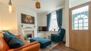 Un salon comprenant un canapé en cuir, un fauteuil turquoise et un pouf, une cheminée, un téléviseur et une grande fenêtre avec des rideaux.