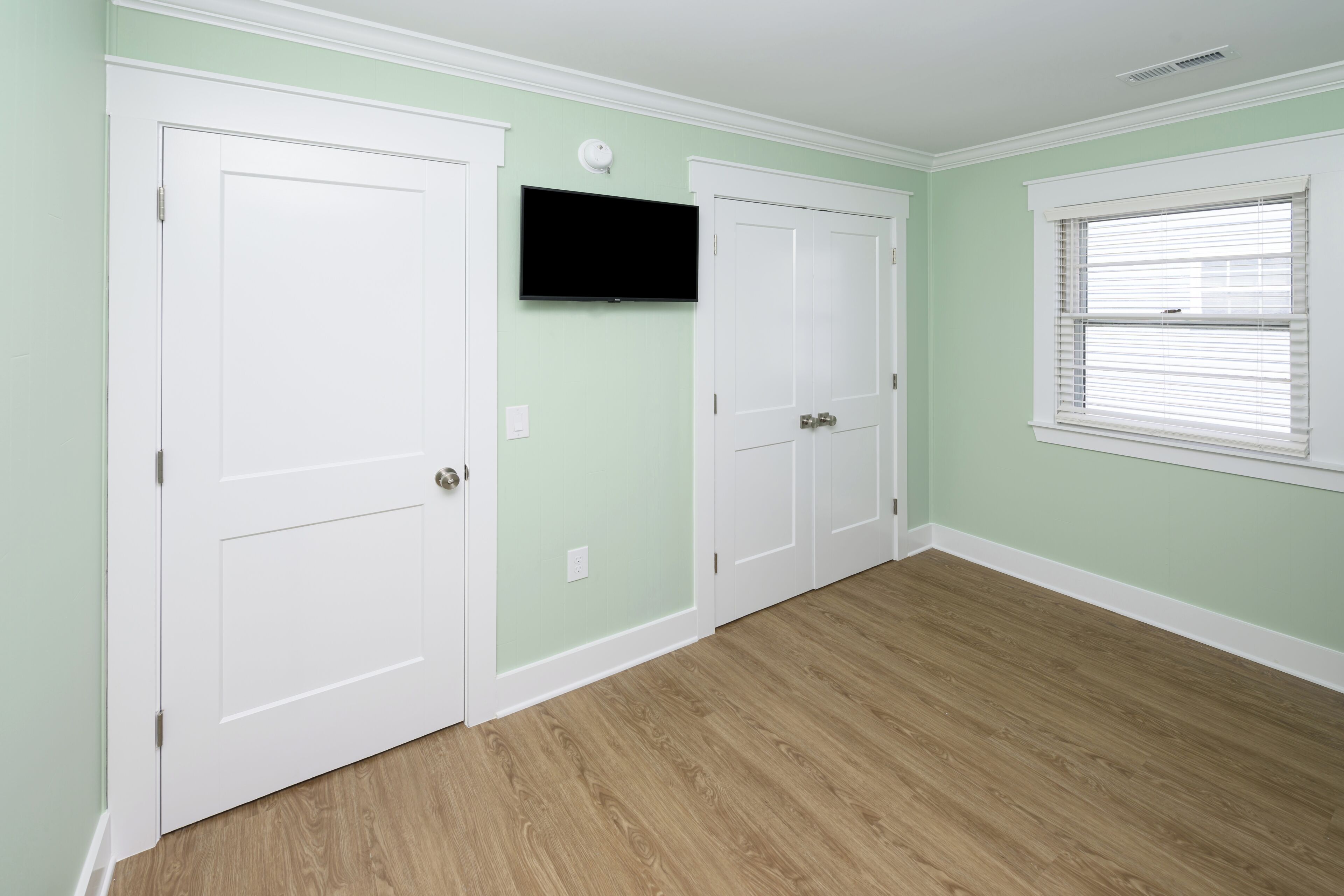 Um quarto com piso de madeira, televisão fixada na parede e portas brancas.