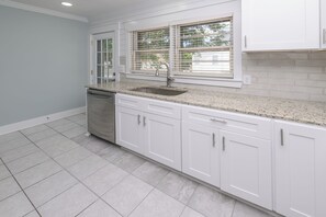 Una cocina con armarios blancos, encimera de granito, pileta de acero inoxidable y ventana con persianas.